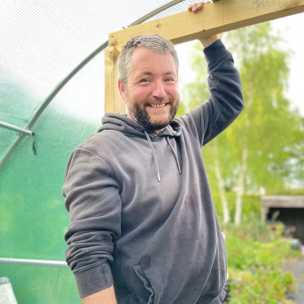 Man in a gray hoodie standing in front of a greenhouse with plants in the background